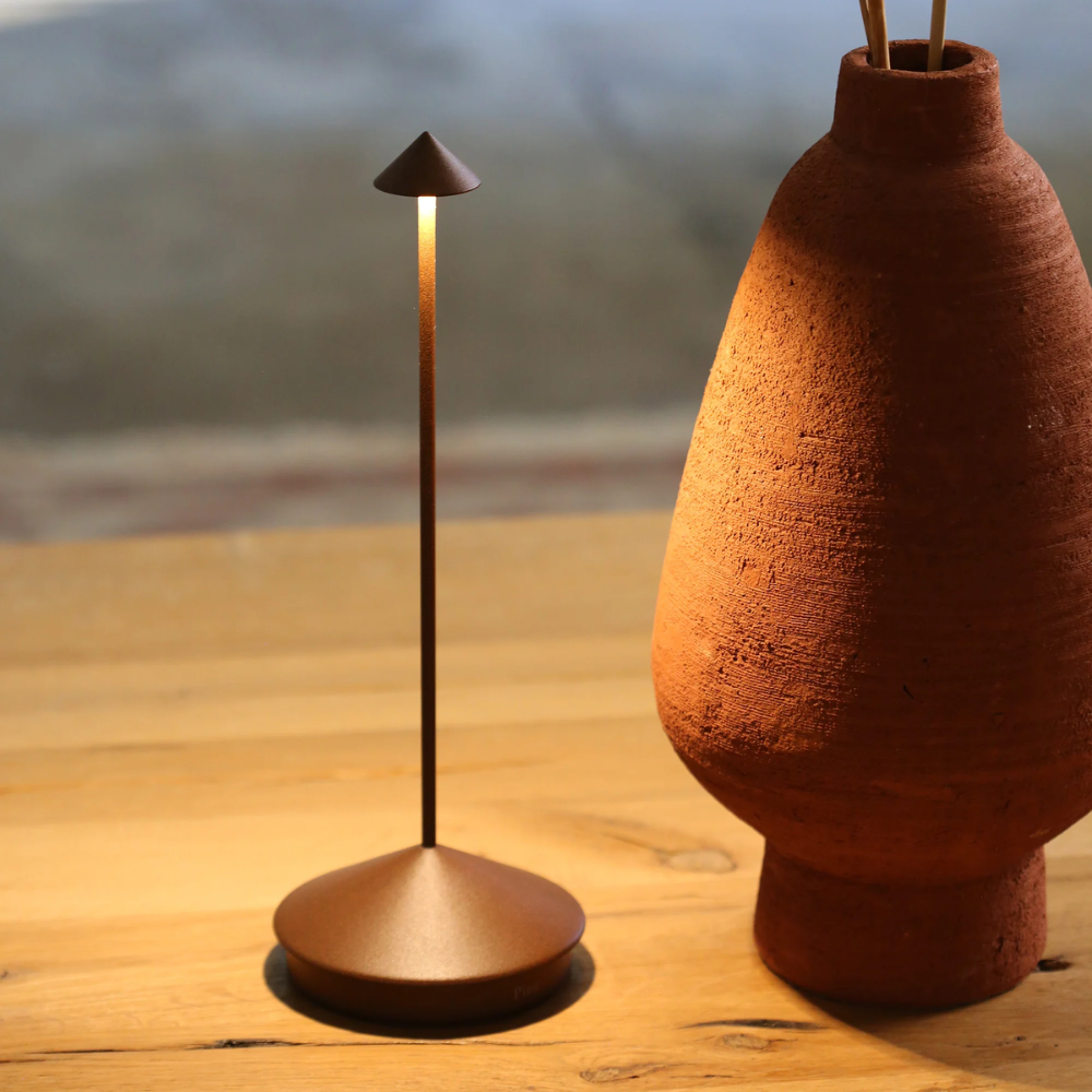 Slim rust-coloured metal table lamp with conical base and cone-shaped shade, next to terracotta vase on table of
