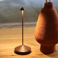 Slim rust-coloured metal table lamp with conical base and cone-shaped shade, next to terracotta vase on table of