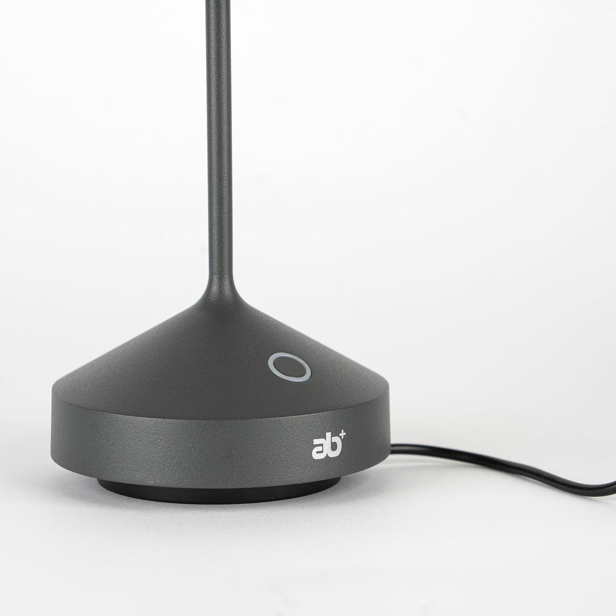 Base con stelo nero opaco, forma conica con parte superiore piatta e logo "ab+" bianco, filo nero su fondo bianco.