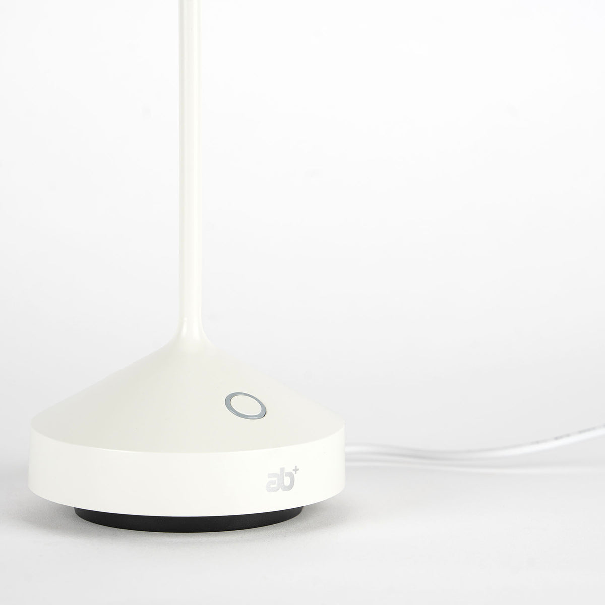 Base bianca di lampada con interruttore circolare grigio, stemma "ab+" e cavo di alimentazione su sfondo chiaro.