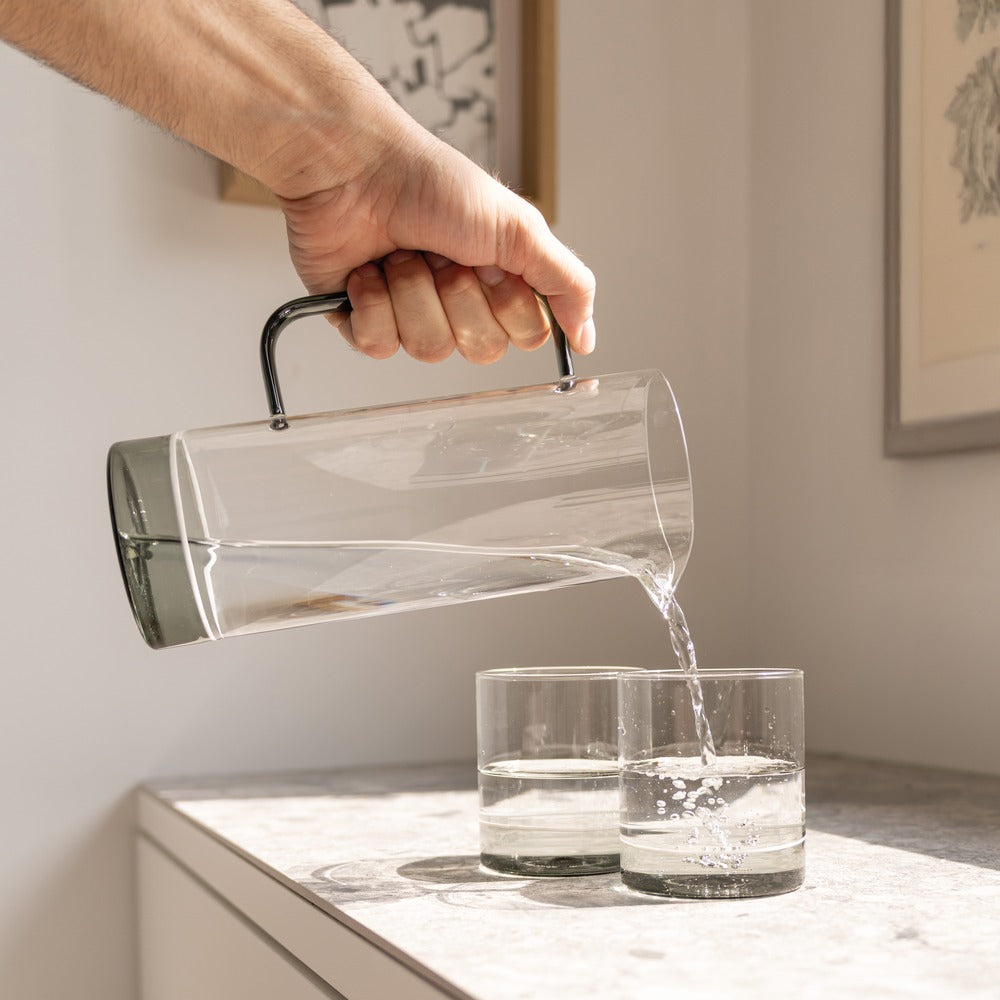 Transparente Glaskaraffe mit schwarzem Griff, gießt Wasser in zwei transparente Gläser auf hellem Tisch. Im Hintergrund ist ein Gemälde zu sehen.