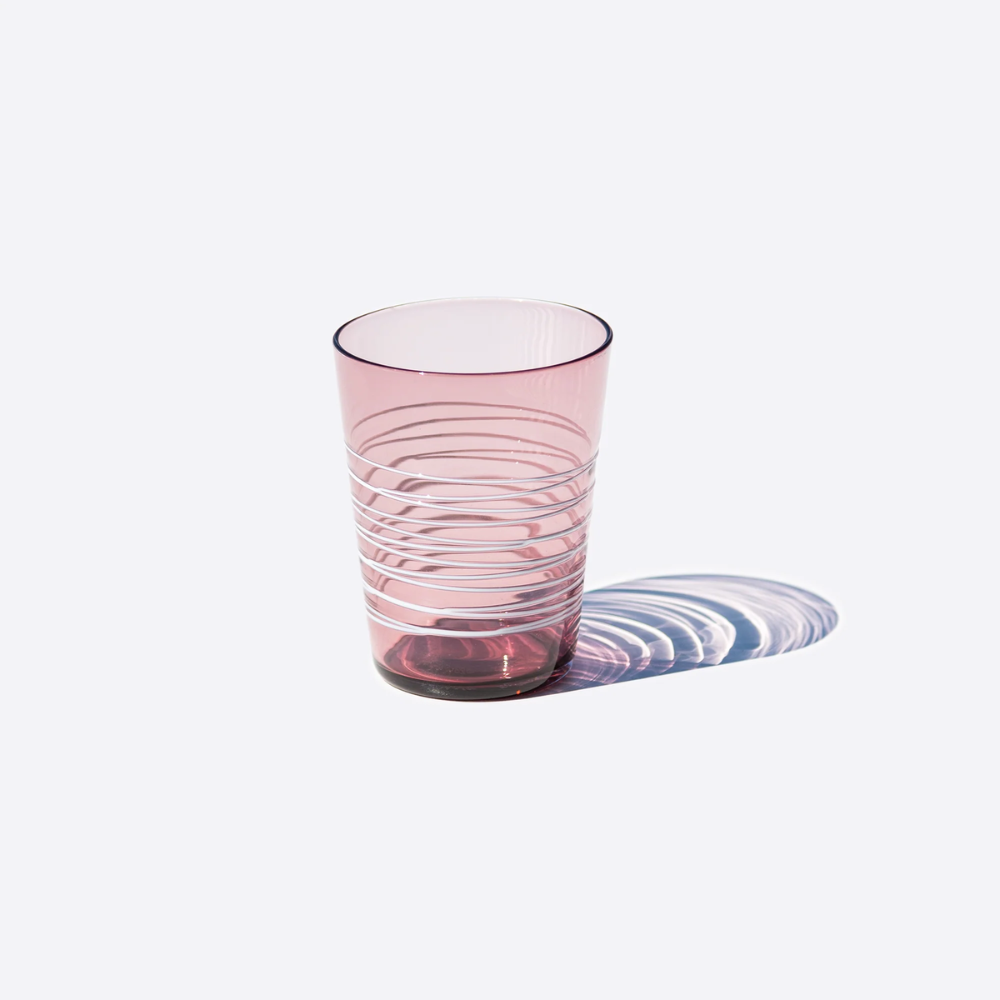 Bicchiere rosa trasparente con decorazione a righe orizzontali bianche su sfondo chiaro con ombra riflessa a destra.