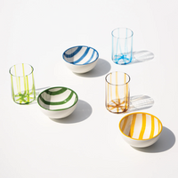Coppette e bicchieri trasparenti con righe colorate rispettivamente in ceramica e vetro, su sfondo bianco con luce naturale.