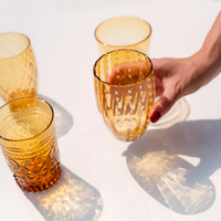 Four amber glass glasses with different decorations, one with white dots held by a hand with red nail polish on white background.