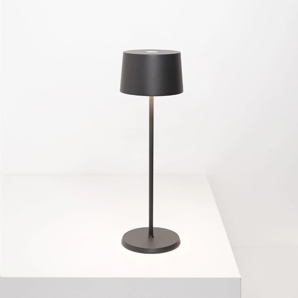 Lampada da tavolo nera in metallo con paralume cilindrico, base piatta rotonda e asta sottile, su superficie bianca minimale.