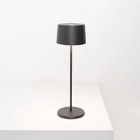 Lampada da tavolo nera in metallo con paralume cilindrico, base piatta rotonda e asta sottile, su superficie bianca minimale.
