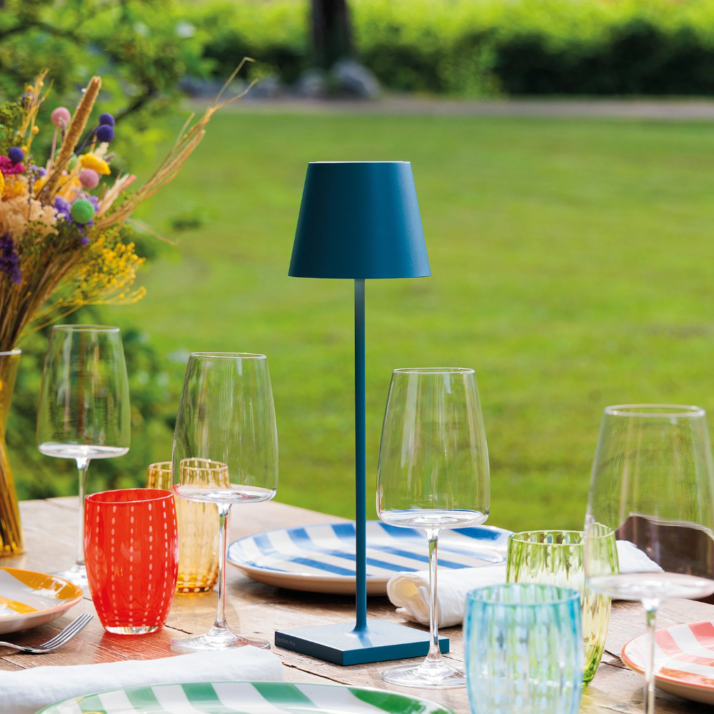 Blue metal table lamp with cylindrical shade on outdoor set table with glasses, colorful plates and flower vase
