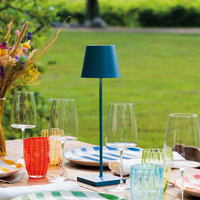 Blue metal table lamp with cylindrical shade on outdoor set table with glasses, colorful plates and flower vase