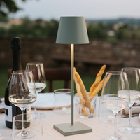 Sage green table lamp with conical shade on square base, wine glasses, bottle and breadsticks on set table