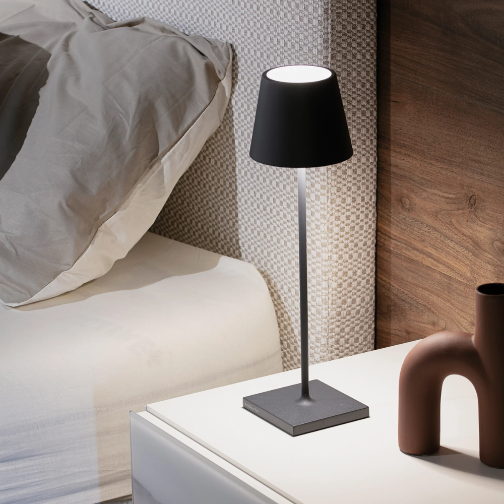 Black metal table lamp with conical shade, lit on white nightstand next to bed with beige bedspread and textile headboard.