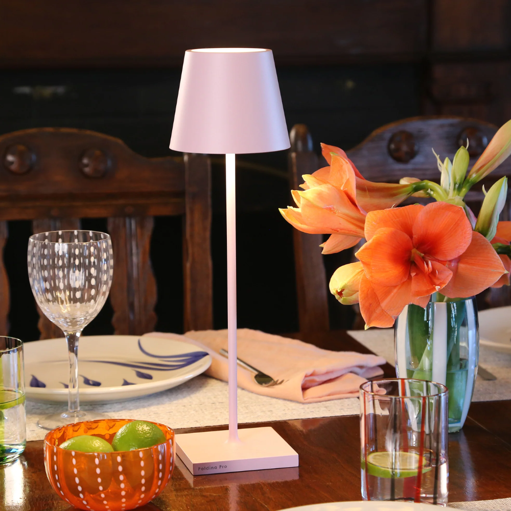 Pink table lamp with square base lit, transparent glass vase with orange flowers, glasses and orange bowl with limes