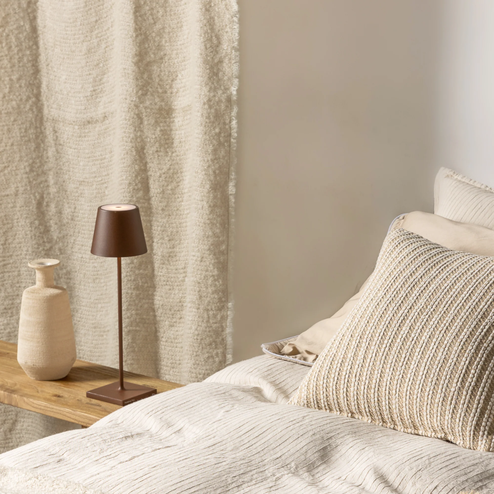 Matte brown metal table lamp with beige fabric vase on wood shelf next to bed with pillows and light beige blanket.