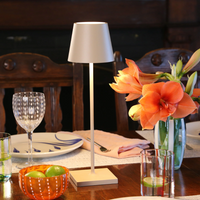 White metal table lamp with conical shade, glass vase with orange flowers, glasses and plates on dark wood table