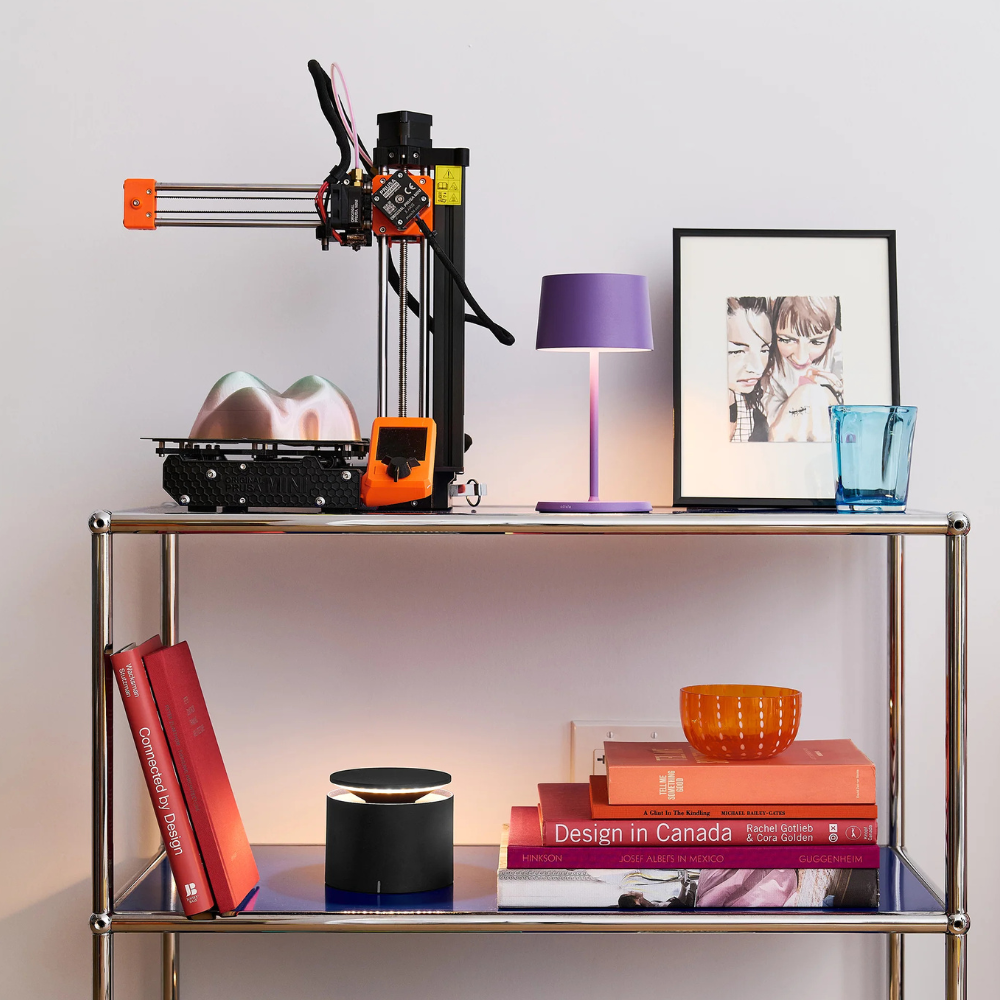 Stampante 3D nera e arancione, lampada viola, cornice con disegno di due persone, bicchiere blu su una mensola metallica con libri rossi e