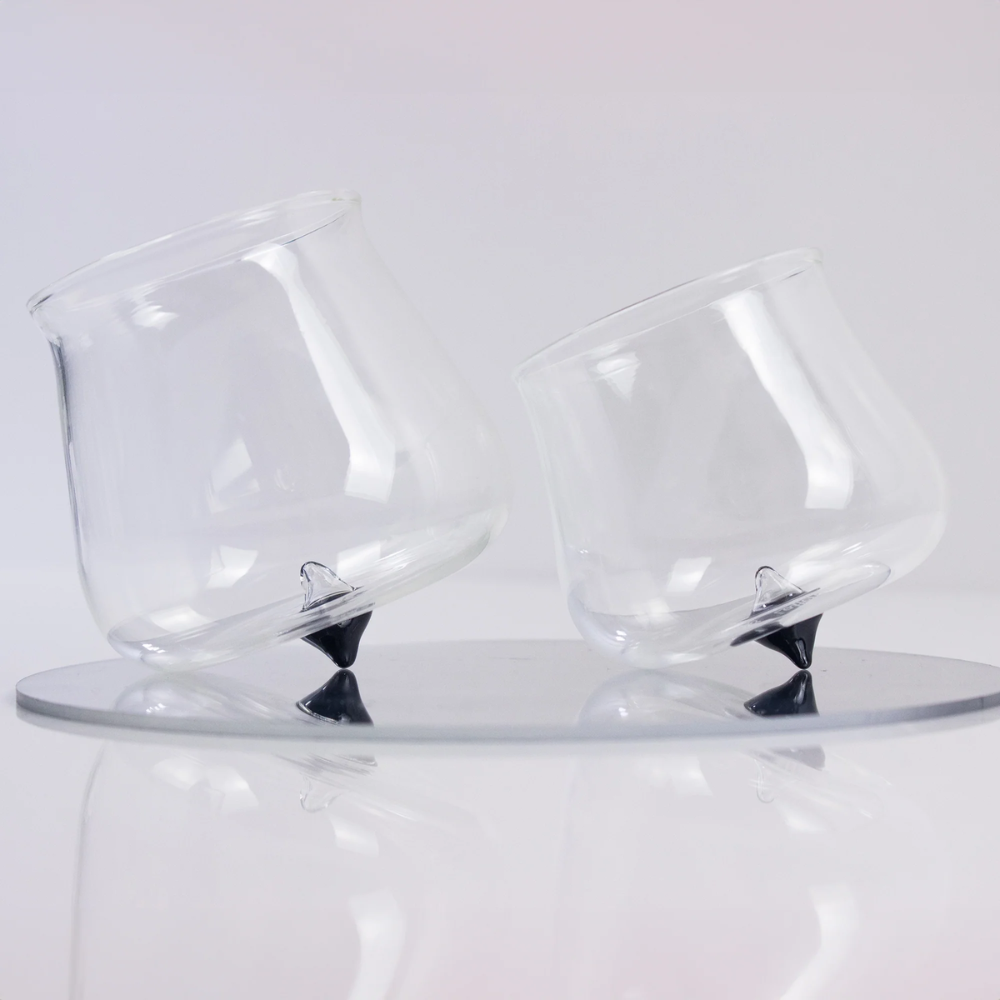 two transparent glass glasses with black conical base, tilted on round reflective glass tray, white background