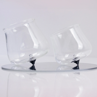 two transparent glass glasses with black conical base, tilted on round reflective glass tray, white background