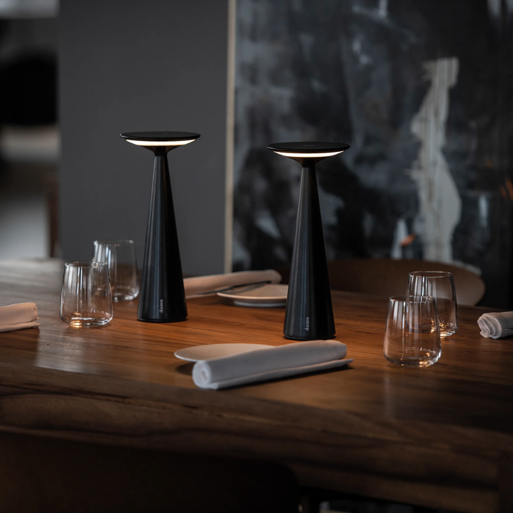 Zwei schwarze konische Tischlampen mit warmem Licht, auf Holztisch mit transparenten Gläsern und gerollten weißen Servietten.