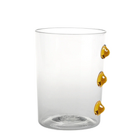 Verre transparent avec trois gouttes décoratives en verre couleur ambre disposées verticalement sur un côté, fond blanc.