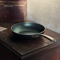 Round black frying pan with silver metal handle, placed on a dark wood surface in a setting with light gray wall.