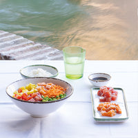 Ciotola grande bianca con riso, verdure e pesce crudo, accompagnata da piatti di sashimi e bicchiere verde, su tavolo vicino a un acquario.