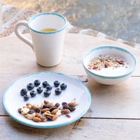 Tazza bianca con bordo azzurro, piatto con mirtilli e mix di frutta secca, ciotola di yogurt con granola, su tavolo di legno.