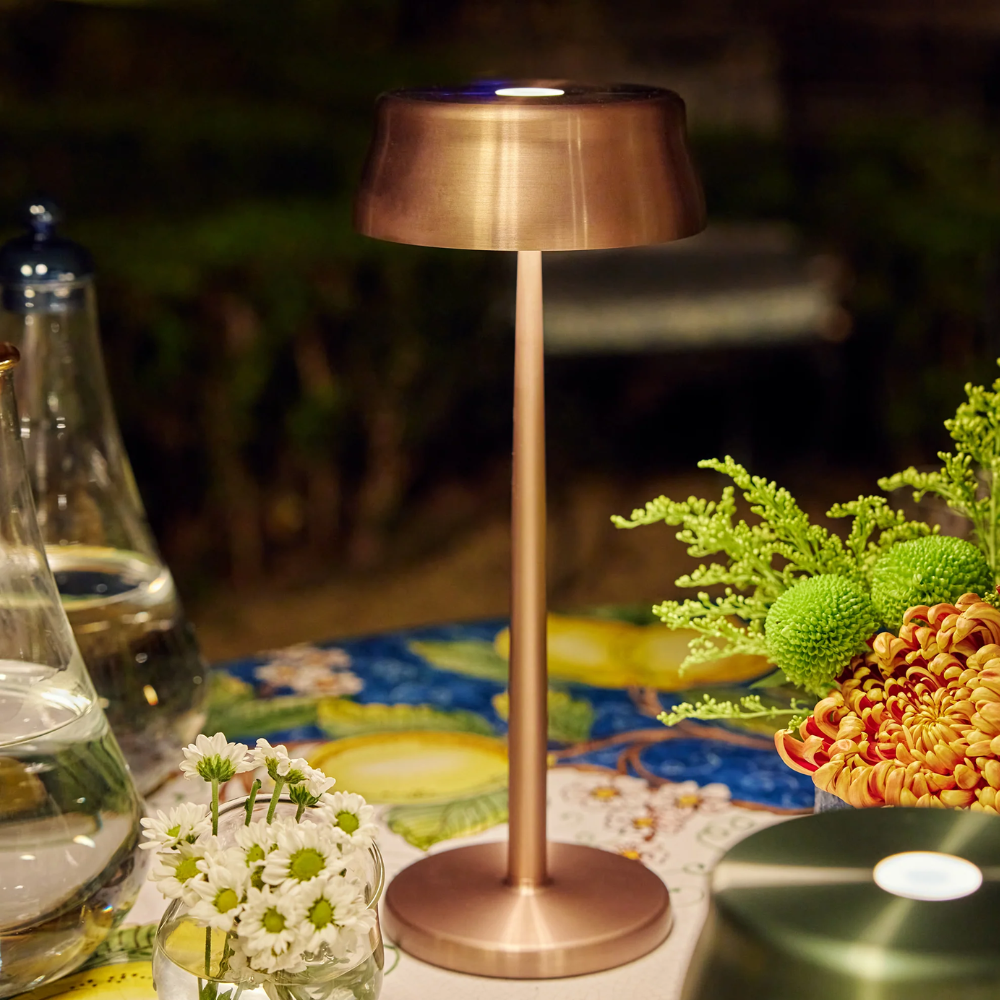 Lampe de table couleur cuivre en métal avec base ronde, entourée de fleurs blanches, orange et vertes sur table décorée.