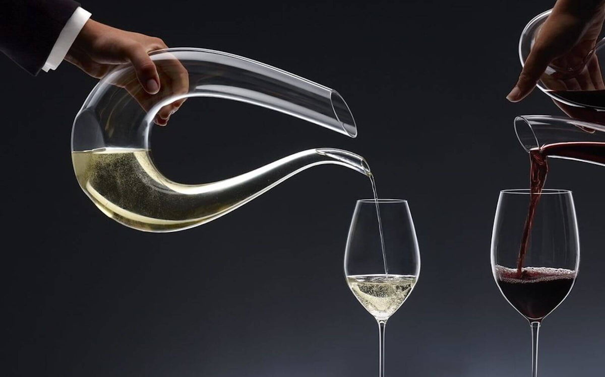 Hand pouring white wine from transparent glass decanter into goblet, other hand pouring red wine into goblet