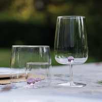 due bicchieri trasparenti in vetro, uno tumbler con tre sfere rosa sul fondo e uno calice con sfera rosa, su tovaglia bianca all'aperto