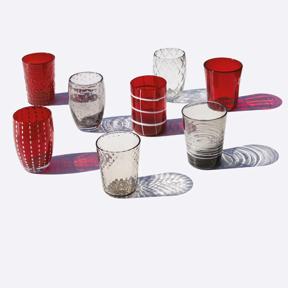 Eight transparent and red glass cups with various textures and patterns, placed on a white surface with colored shadows.
