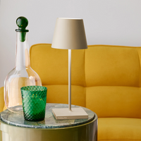 Lampada da tavolo beige con paralume con base quadrata accanto a bottiglia trasparente con tappo verde e bicchiere verde ricoperti da un
