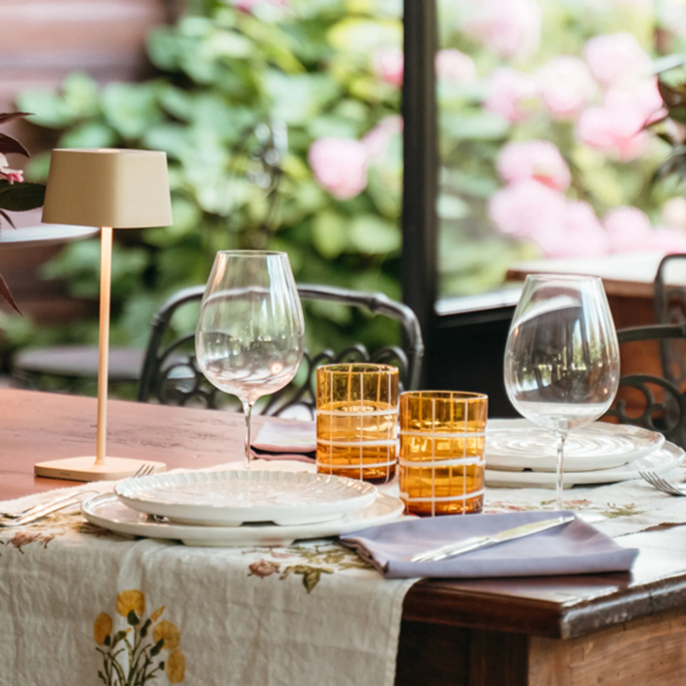 Wood table with floral white tablecloth, two transparent wine glasses, two orange glasses, white plates and beige lamp