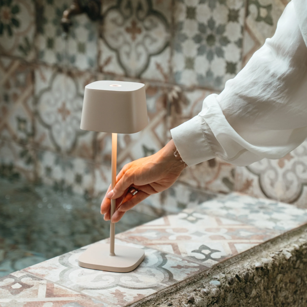 Beige table lamp with square base, slim stem and square shade, lit near pool edge with decorated tiles, hand