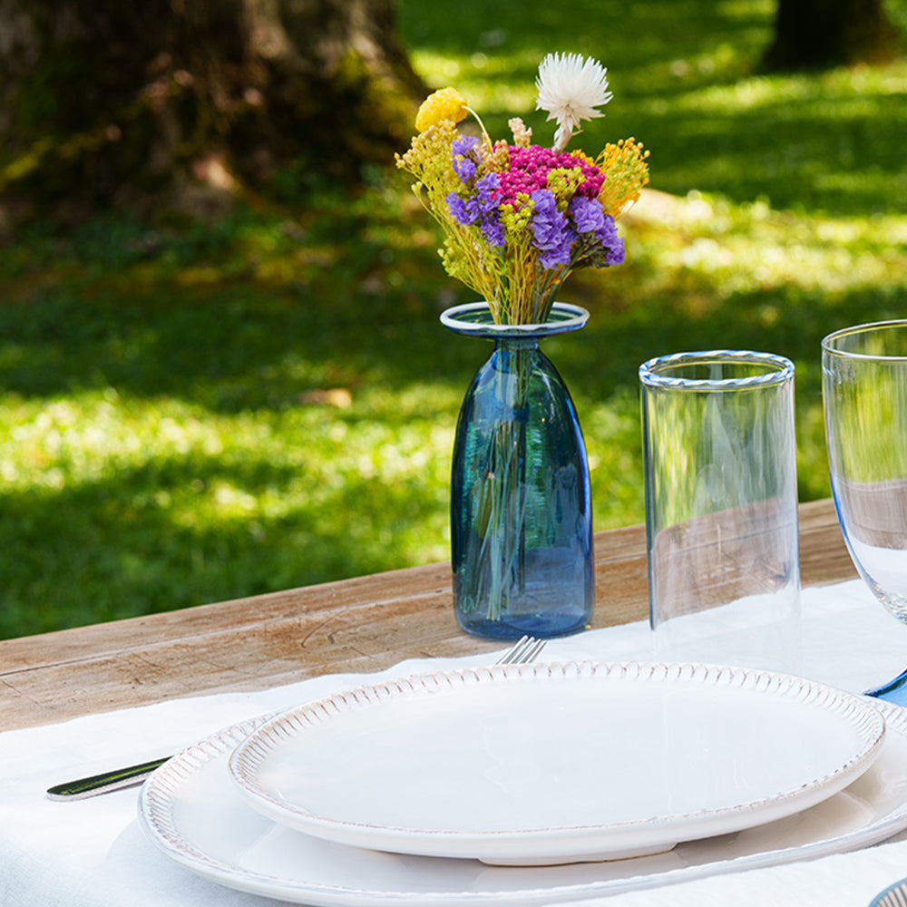 Blue glass vases with colorful flowers, white plates and transparent glasses on a wooden table in a green sunny setting.