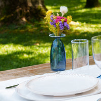 Blue glass vases with colorful flowers, white plates and transparent glasses on a wooden table in a green sunny setting.