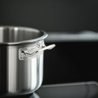 Glanzende roestvrijstalen pan met korte steel, keukenomgeving met wazige zwarte kookplaat op de achtergrond.
