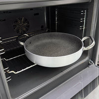 Ronde koekenpan met twee glanzende metalen handgrepen en donker anti-aanbak interieur in open grijze oven.