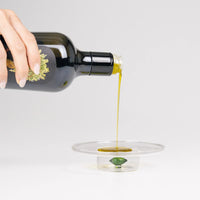 Bottiglia nera di olio d'oliva, versata su un piatto trasparente, con olio giallo brillante che scorre, sfondo grigio chiaro.