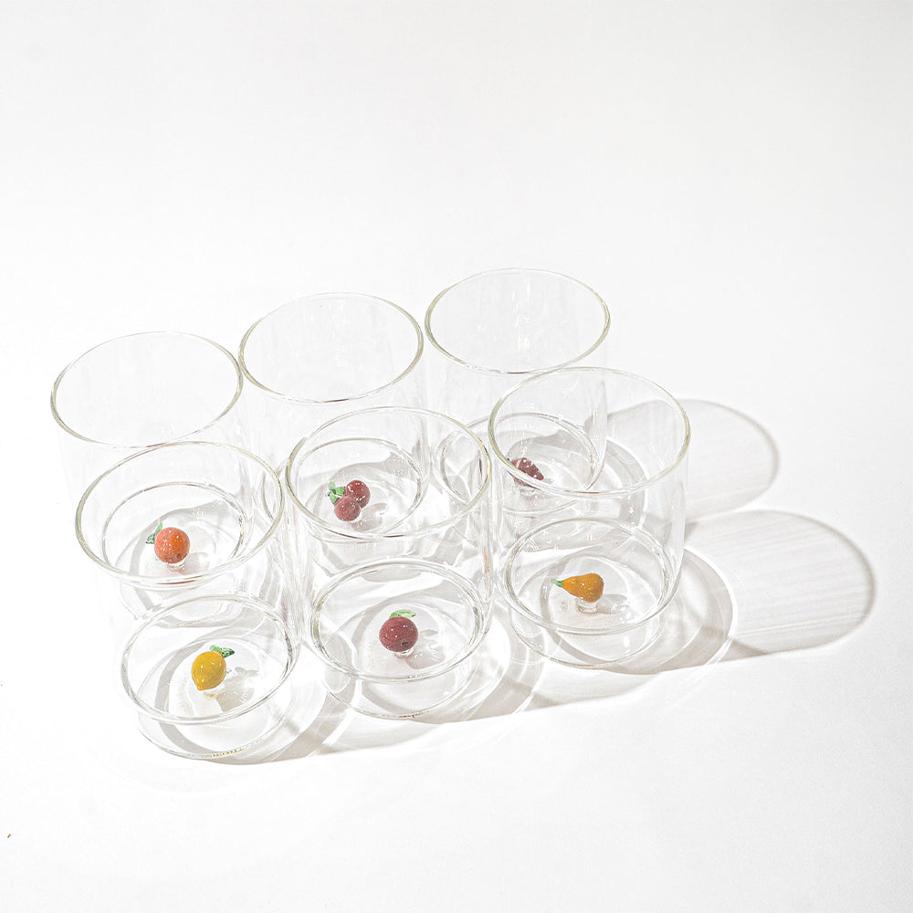 Set di otto bicchieri in vetro trasparente con diversi piccoli frutti colorati decorativi incastonati alla base, su sfondo bianco.