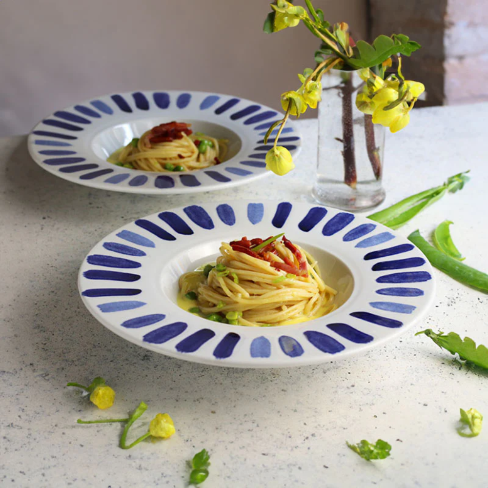 deux assiettes blanches à bord décoré de coups de pinceau bleus contenant des spaghettis aux petits pois et tomates cerises sur table claire avec vase en verre et