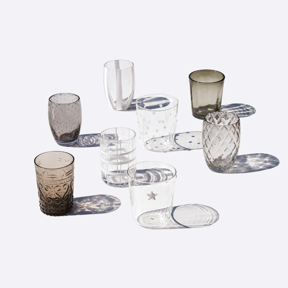 Set of eight transparent and smoked glass cups with various decorations such as polka dots, stripes, engravings and star patterns, on a light background.