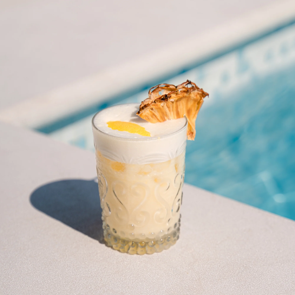 Decorated glass tumbler with creamy yellow drink, white foam, dried pineapple slice and lemon zest, poolside.