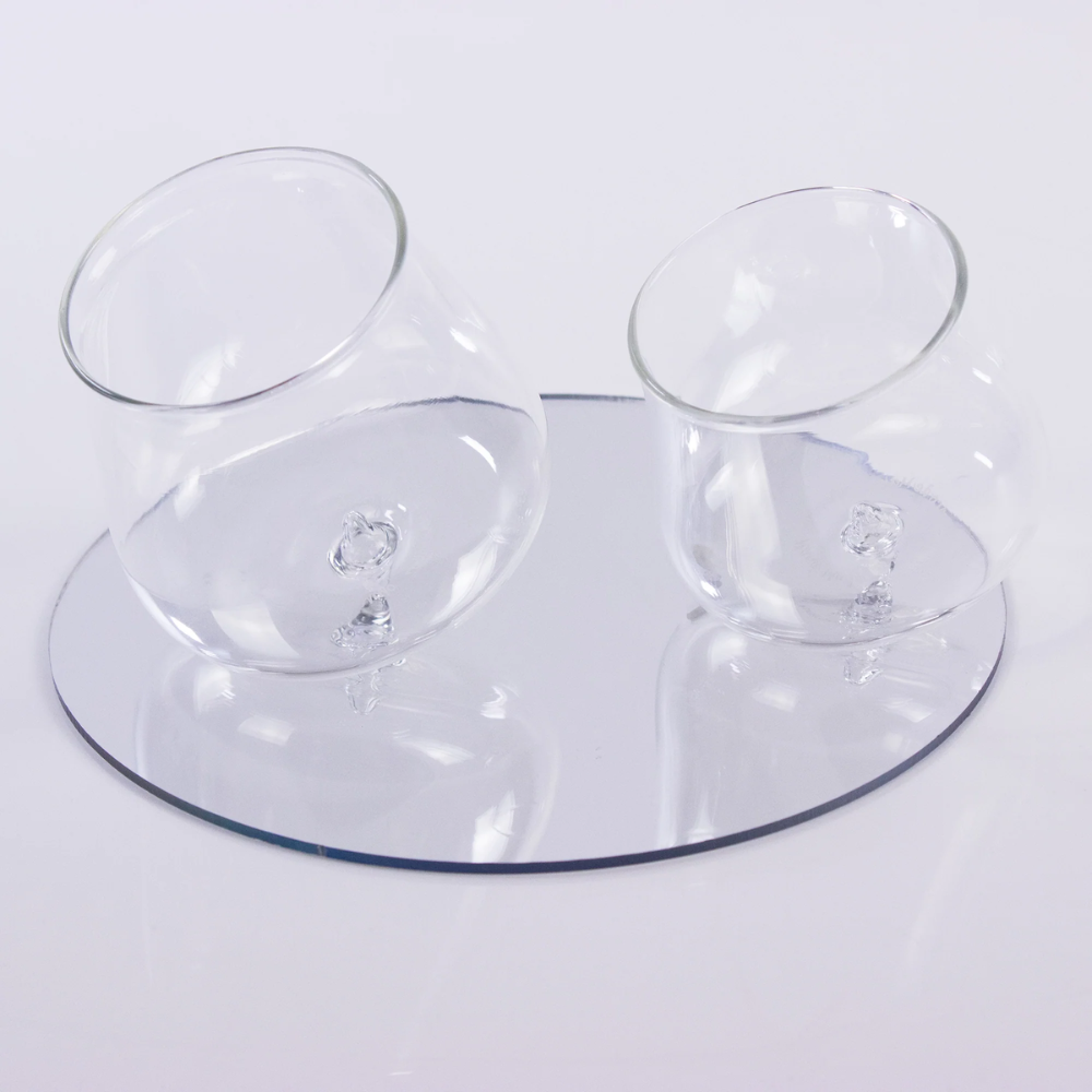 two transparent glass glasses tilted on round base with reflective surface, simple white background