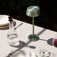 Lampe de table verte en métal avec base circulaire, sur nappe beige avec bouteille d'eau, verre, assiettes et fourchettes.