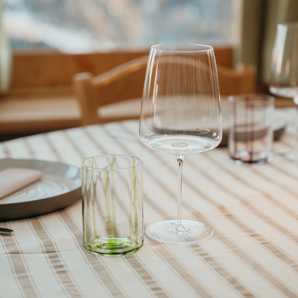 bicchiere verde trasparente cilindrico e calice trasparente con gambo su tovaglia beige a righe sottili in ambiente interno con tavolo