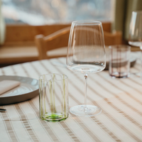 bicchiere verde trasparente cilindrico e calice trasparente con gambo su tovaglia beige a righe sottili in ambiente interno con tavolo