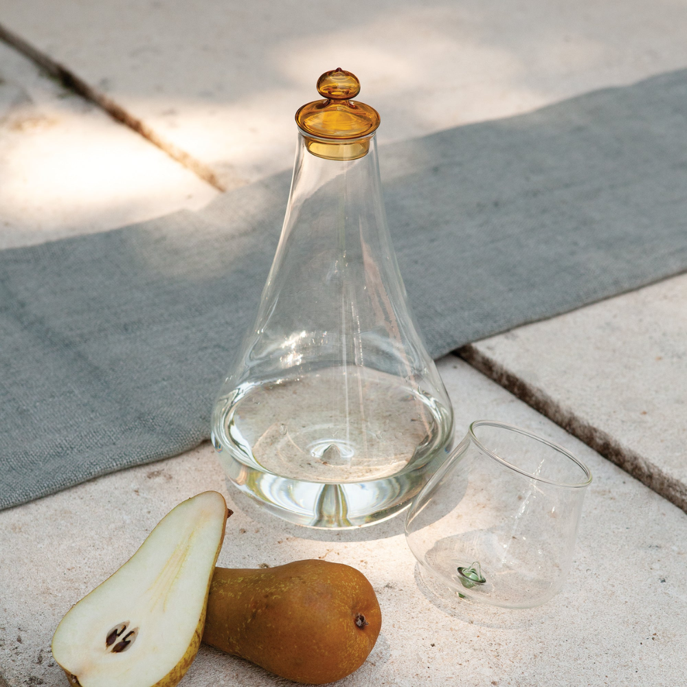Transparent glass decanter and glass with amber cap, a whole pear and a sliced one, on light stone surface with grey cloth