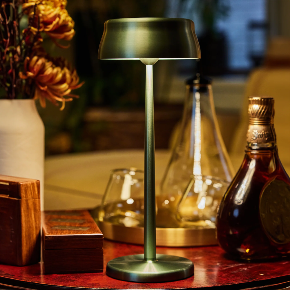 Lampe verte en métal avec base circulaire, allumée sur table rouge avec vase de fleurs séchées, bouteille et contenants en bois.