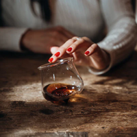 Female hand with red nails placing upside-down transparent glass with amber liquid on dark wood table