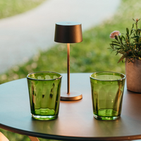 Two green glass glasses, a bronze table lamp and a vase with flowers and leaves on round outdoor table.