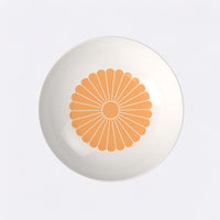 Piatto bianco con decorazione centrale arancione a forma di fiore stilizzato. Sfondo chiaro.